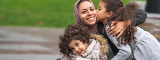 A family of Middle Eastern descent is spending time together. A young mother is playing at the park with her two young daughters. The mother is kneeling down and embracing her children. The happy group is smiling. The older daughter is giving her mom a kiss on the cheek.